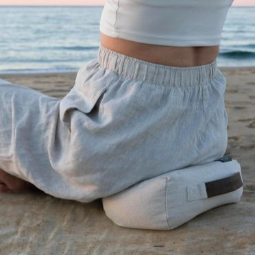 Osoba w lnianych spodniach i białym topie siedzi na beżowej poduszce do medytacji na piasku, w tle spokojne morze i niebo o zachodzie słońca.